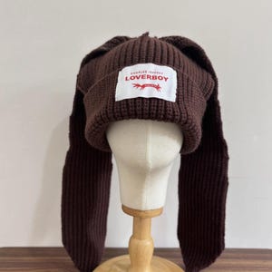 May include: A brown knit hat with long ear-like flaps. The hat features a rectangular white label with the text "CHARLES JEFFREY LOVERBOY" in black and red. The hat is displayed on a mannequin head.