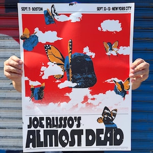 May include: A red and white poster featuring the text "JOE RUSSO'S ALMOST DEAD" in bold black letters. The poster has a red background with white clouds and illustrations of butterflies. The poster also includes the dates and locations of the event.