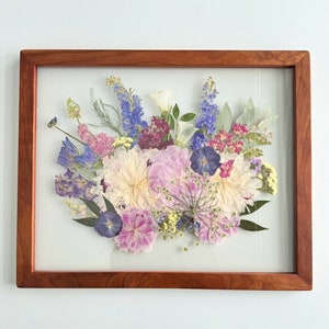 以下が含まれることがあります： ピンク、白、青、紫の色合いのさまざまな花を使った、額装された押し花のアレンジメント。花はブーケの形にアレンジされ、2枚のガラス板で挟まれています。フレームは木製の自然な仕上げです。