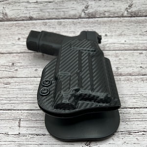 May include: A black carbon fiber gun holster with a black plastic clip. The holster has a textured surface and a black button on the side.