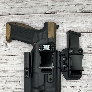 May include: A black leather holster with a magazine carrier for a handgun. The holster has a belt clip and adjustable retention. The handgun is a Canik TP9SFx with a brown and black finish.