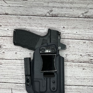May include: A black polymer holster with a clip for carrying a handgun. The holster has a textured surface and a black metal clip.