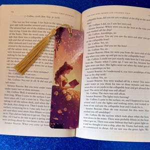 May include: Open book with a decorative bookmark. The bookmark features a princess illustration in a yellow gown, holding a book. A gold tassel hangs from the top. The book pages are filled with text.