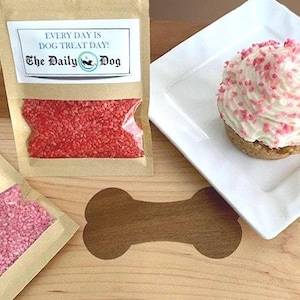 May include: Two small bags of pink and red sprinkles, labeled "The Daily Dog" with the text "Every day is dog treat day!"  A white cupcake with pink sprinkles on top is on a white plate.
