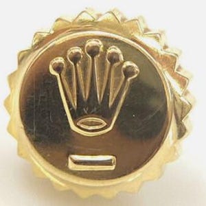May include: A gold-colored watch crown with a five-pointed crown logo and a horizontal line below it.