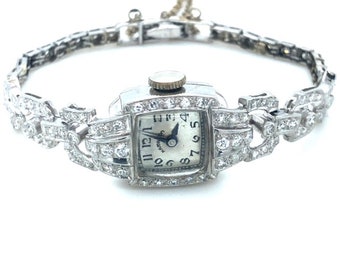 May include: A vintage wristwatch with a square face and diamond-encrusted band. The watch face is white with black Roman numeral hour markers and black hands. The watch is set in a silver-toned metal.