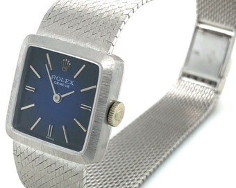 May include: A silver Rolex Geneve wristwatch with a square blue face and a silver mesh band.