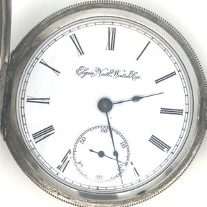 Könnte beinhalten: Eine silberne Taschenuhr mit römischen Ziffern für die Stunden und einem weißen Zifferblatt. Auf dem Zifferblatt der Uhr steht der Text "Elgin Natl. Watch Co.".