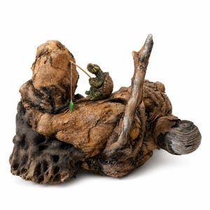 May include: A small green turtle figurine holding a thin wooden fishing rod with a green lure, positioned on a large piece of textured, weathered brown driftwood that features natural knots, crevices, and a smooth, rounded end section.