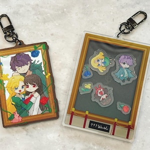 Può includere: Due portachiavi con personaggi anime. Il primo portachiavi è una cornice di legno con un'immagine di tre personaggi. Il secondo portachiavi è una cornice trasparente con un'immagine di quattro personaggi. Il testo sul secondo portachiavi recita "??? World".