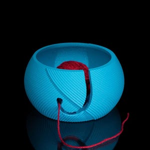 May include: A blue yarn bowl with a red ball of yarn inside. The yarn is threaded through a hole in the bowl.