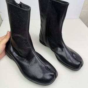 May include: A pair of black leather boots with a split-toe design. The boots have a mid-calf height and a zipper on the side. The boots are made of a smooth, shiny leather and have a flat sole.