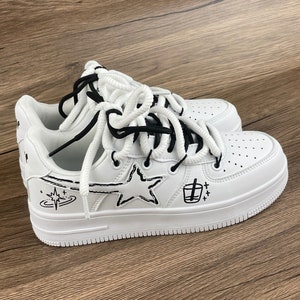 May include: White leather sneakers with black doodle designs including a star, a planet, and a cup. The laces are white with black accents.