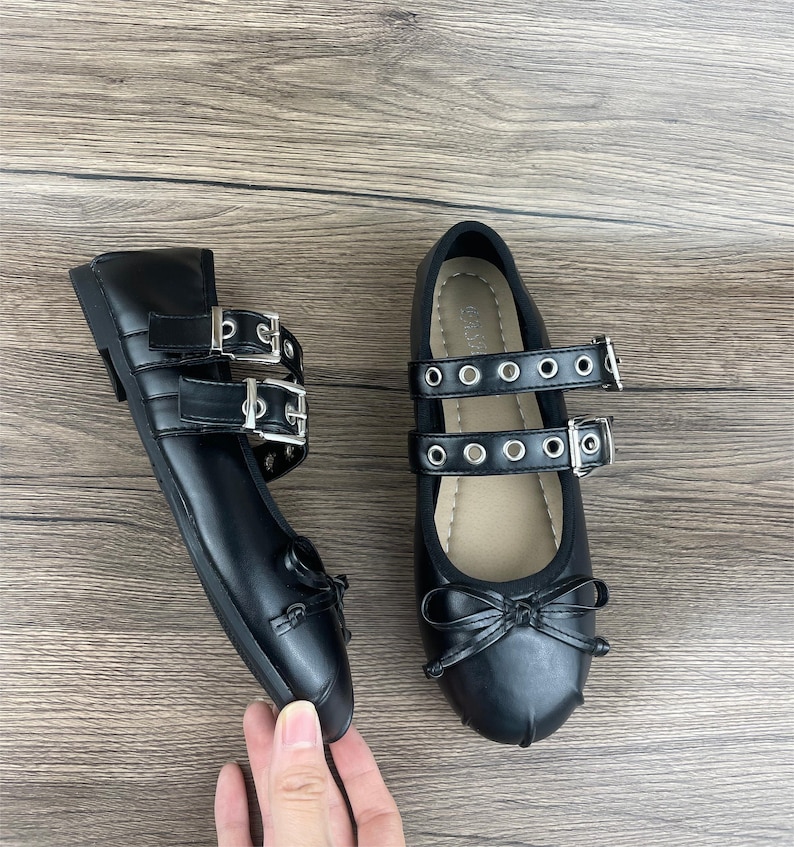 May include: A pair of black Mary Jane style flats with double straps and silver buckles. The shoes have a small bow detail on the toe and are made of a smooth, shiny material. The shoes are displayed on a wooden surface.