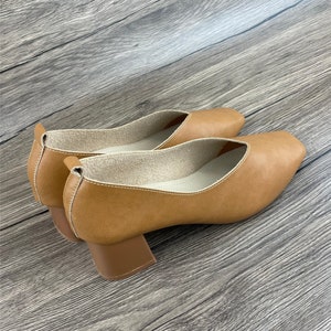 May include: A pair of brown leather heeled shoes with a square toe. The shoes have a block heel and a white lining.