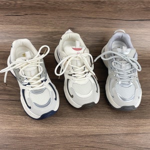 May include: Three pairs of white trainers with grey and silver accents. The trainers have white laces and a chunky sole.