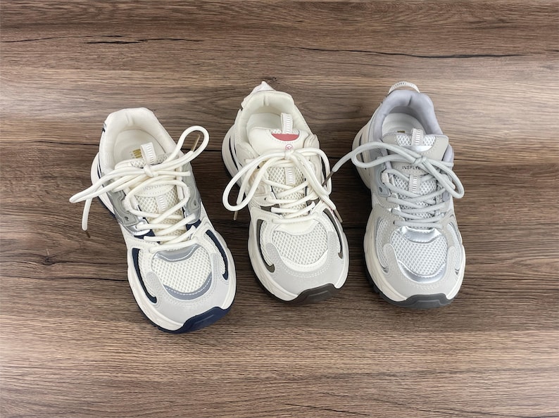 May include: Three pairs of white trainers with grey and silver accents. The trainers have white laces and a chunky sole.