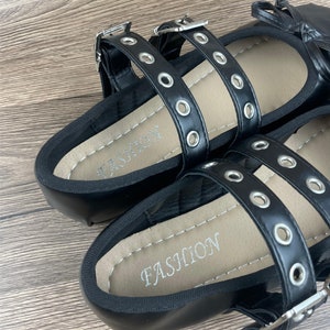 May include: Black flat sandals with multiple straps and silver-tone buckles. The straps are adorned with silver eyelets. The footbed is tan with the word "FASHION" printed in silver. The sandals are on a wooden surface.