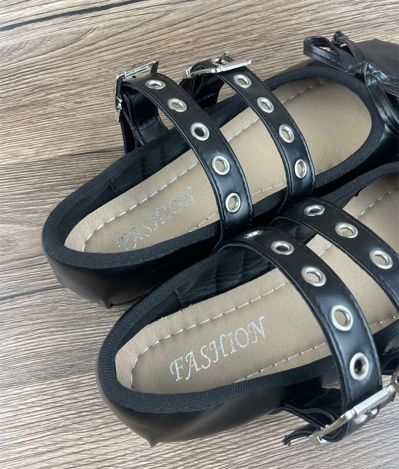May include: Black flat sandals with multiple straps and silver-tone buckles. The straps are adorned with silver eyelets. The footbed is tan with the word "FASHION" printed in silver. The sandals are on a wooden surface.