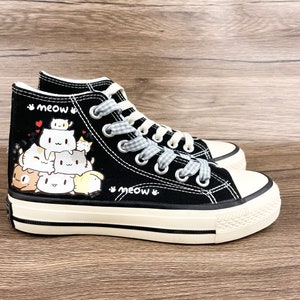 May include: Black high-top canvas sneakers with a white sole and a design of stacked cartoon cats. The laces are white with a black and white checkered pattern. The words "meow" are written on the side of the shoe.