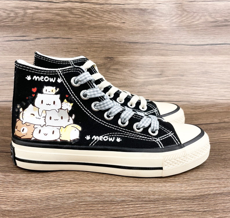 May include: Black high-top canvas sneakers with a white sole and a design of stacked cartoon cats. The laces are white with a black and white checkered pattern. The words "meow" are written on the side of the shoe.