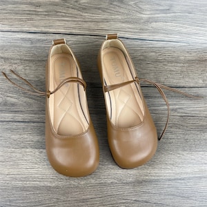 May include: A pair of brown leather flats with a tie closure. The shoes have a rounded toe and a flat heel. The brand name "TOMU" is printed on the inside of each shoe.