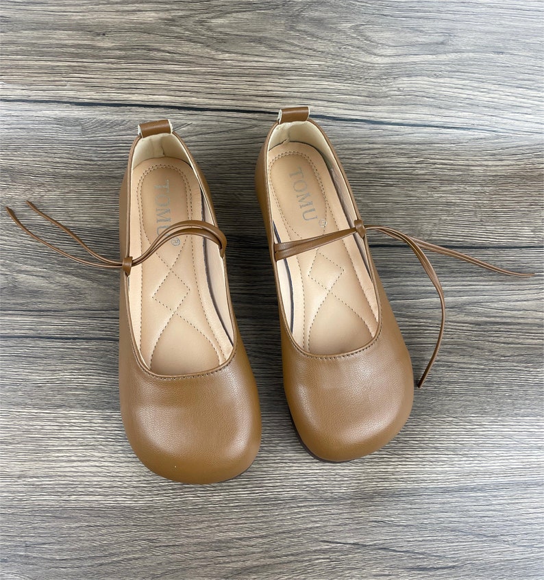 May include: A pair of brown leather flats with a tie closure. The shoes have a rounded toe and a flat heel. The brand name "TOMU" is printed on the inside of each shoe.