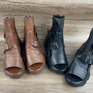 Puede incluir: Cuatro botines de tobillo con punta abierta en marrón y negro. Las botas tienen una suela gruesa, una correa en el pie y un cierre de hebilla. Las botas marrones están a la izquierda y las negras a la derecha.