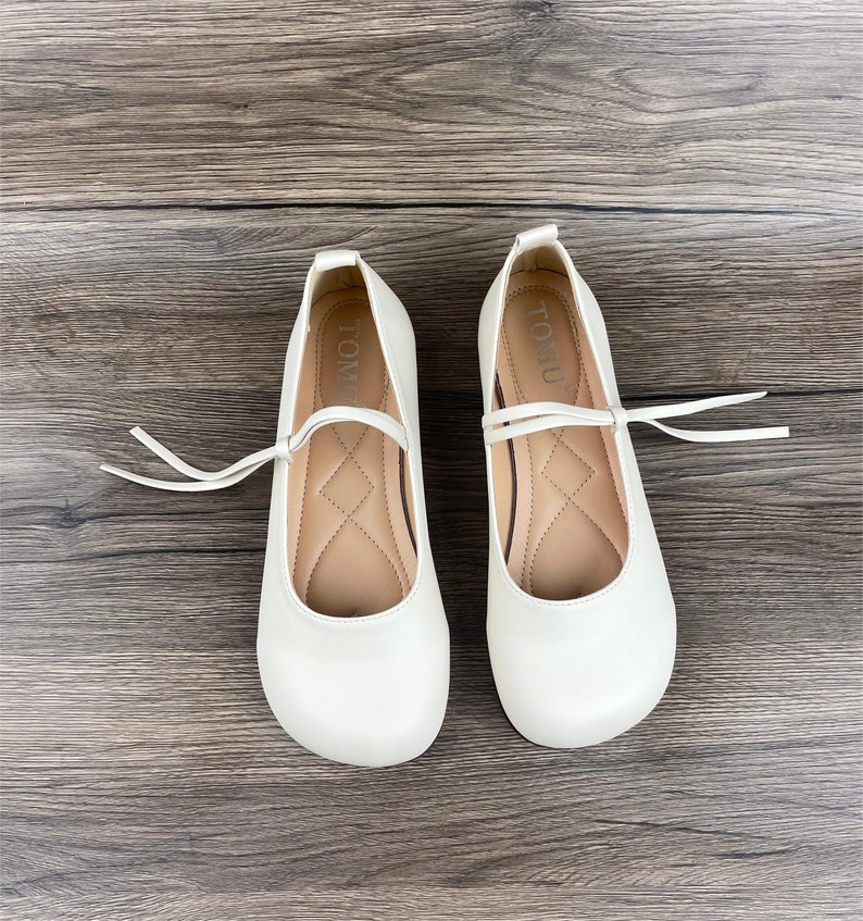 May include: A pair of white leather ballet flats with a small bow and a tie at the ankle. The shoes have the word "TOMU" written on the inside.