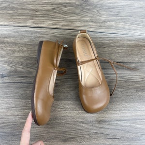 May include: A pair of brown leather flats with a tie closure. The shoes have a rounded toe and a flat heel. The brand name "TOMU" is printed on the inside of the right shoe.