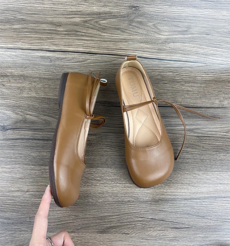 May include: A pair of brown leather flats with a tie closure. The shoes have a rounded toe and a flat heel. The brand name "TOMU" is printed on the inside of the right shoe.