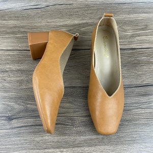 May include: A pair of brown leather heeled shoes with a square toe. The shoes have a block heel and a pointed vamp. The brand name "Elemenday" is printed on the inside of the right shoe.
