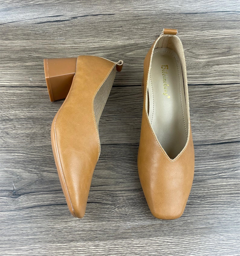 May include: A pair of brown leather heeled shoes with a square toe. The shoes have a block heel and a pointed vamp. The brand name "Elemenday" is printed on the inside of the right shoe.