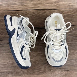 May include: A pair of white trainers with a silver mesh design and a chunky sole. The trainers have a navy blue trim and white laces.