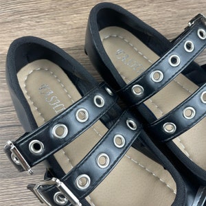 May include: Black Mary Jane style shoes with double straps and silver-tone grommets. The straps have silver buckles. The insole has the word "FASHION" printed on it. The shoes are on a wooden surface.