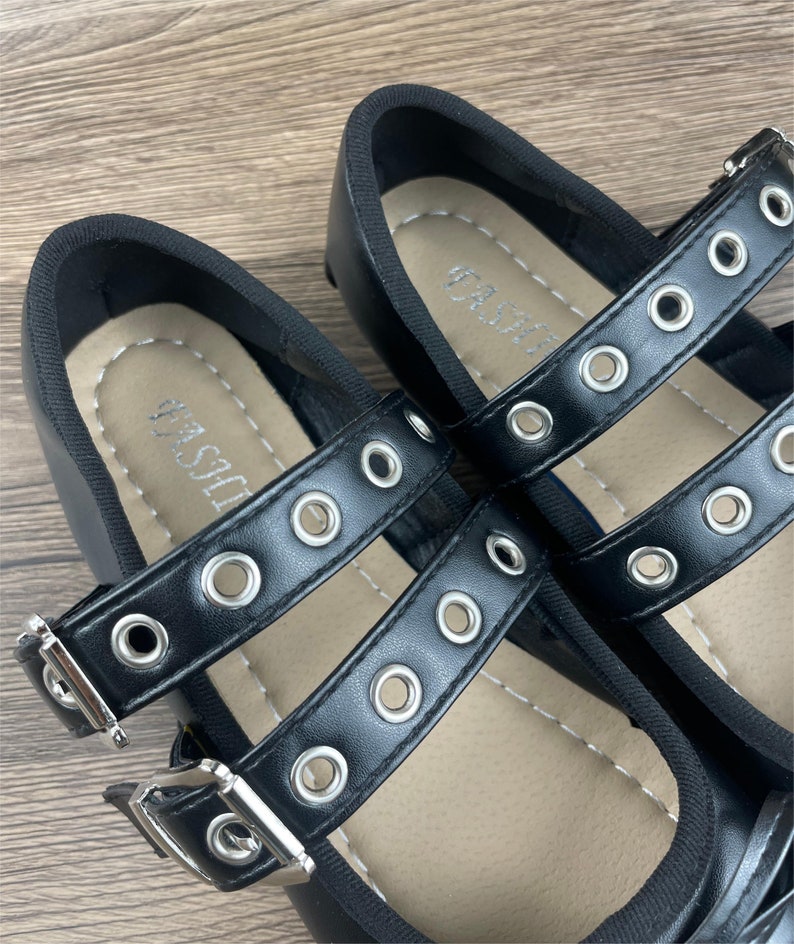 May include: Black Mary Jane style shoes with double straps and silver-tone grommets. The straps have silver buckles. The insole has the word "FASHION" printed on it. The shoes are on a wooden surface.