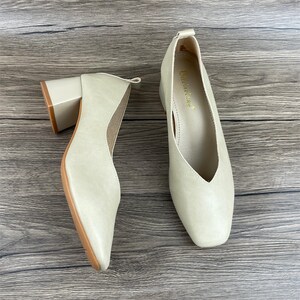 May include: A pair of beige leather heeled shoes with a pointed toe and a v-shaped opening at the front. The shoes have a block heel and are on a wooden surface.