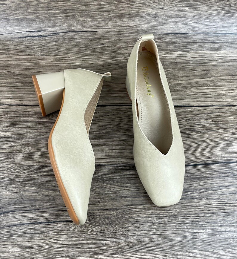 May include: A pair of beige leather heeled shoes with a pointed toe and a v-shaped opening at the front. The shoes have a block heel and are on a wooden surface.