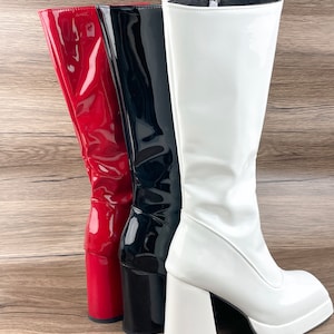 K&ouml;nnte beinhalten: Drei Paar kniehohe Stiefel in Rot, Schwarz und Wei&szlig;. Die Stiefel haben einen dicken Plateauabsatz und eine gl&auml;nzende Oberfl&auml;che.