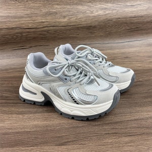 May include: A pair of white and silver trainers with a chunky platform sole. The trainers have a mesh and suede upper with a lace-up closure.