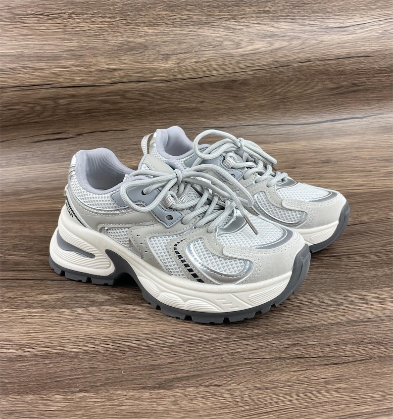May include: A pair of white and silver trainers with a chunky platform sole. The trainers have a mesh and suede upper with a lace-up closure.