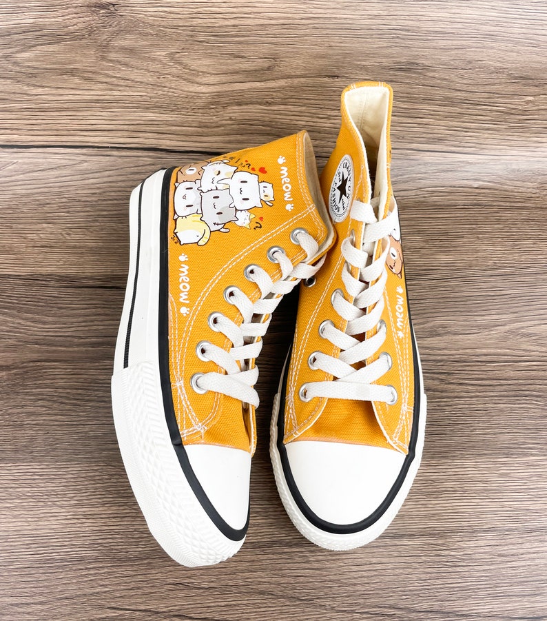 May include: A pair of yellow high-top canvas sneakers with a black trim. The sneakers feature a cartoon illustration of cats with the text "Meow" on each shoe.