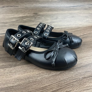 May include: A pair of black ballet flats with double straps, silver buckles, and bows. The shoes have a rounded toe and are made of a smooth material. The background is a wooden surface.