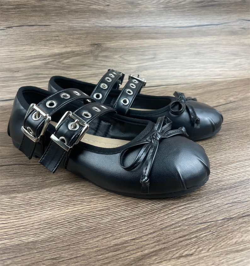 May include: A pair of black ballet flats with double straps, silver buckles, and bows. The shoes have a rounded toe and are made of a smooth material. The background is a wooden surface.