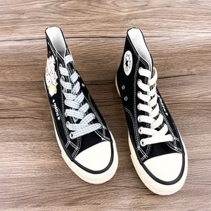 May include: A pair of black high-top canvas sneakers with white laces. One shoe has a blue and white checkered lace and a cartoon cat design on the side. The other shoe has a white star logo on the side.