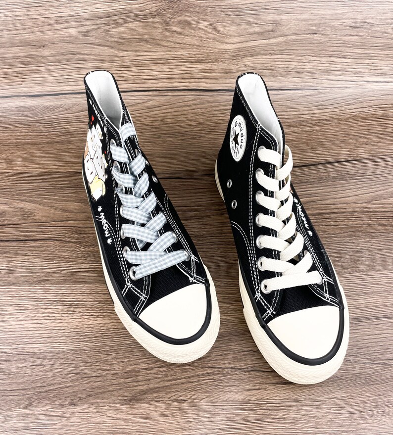 May include: A pair of black high-top canvas sneakers with white laces. One shoe has a blue and white checkered lace and a cartoon cat design on the side. The other shoe has a white star logo on the side.