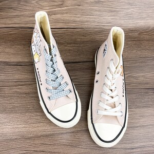 May include: A pair of light beige high-top canvas sneakers with white laces and a black trim. The sneakers feature a cartoon illustration of cats and the text "Meow."