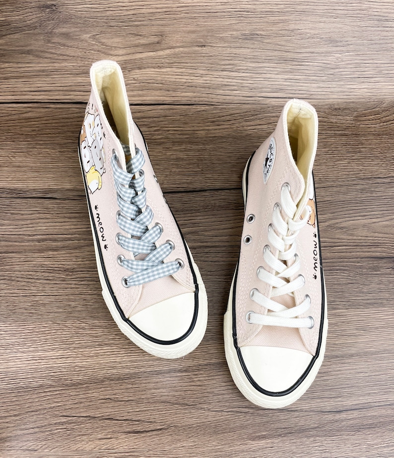May include: A pair of light beige high-top canvas sneakers with white laces and a black trim. The sneakers feature a cartoon illustration of cats and the text "Meow."