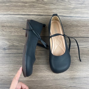 May include: A pair of black leather flats with a rounded toe and a small bow on the front. The shoes have a black ribbon that ties around the ankle. The shoes are on a wooden surface.