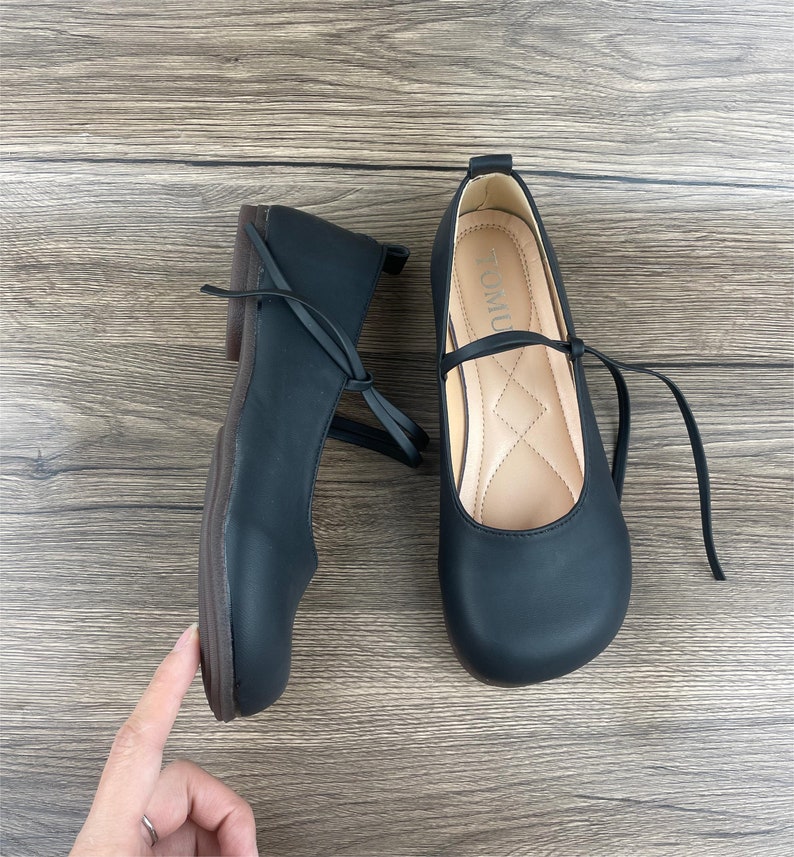 May include: A pair of black leather flats with a rounded toe and a small bow on the front. The shoes have a black ribbon that ties around the ankle. The shoes are on a wooden surface.